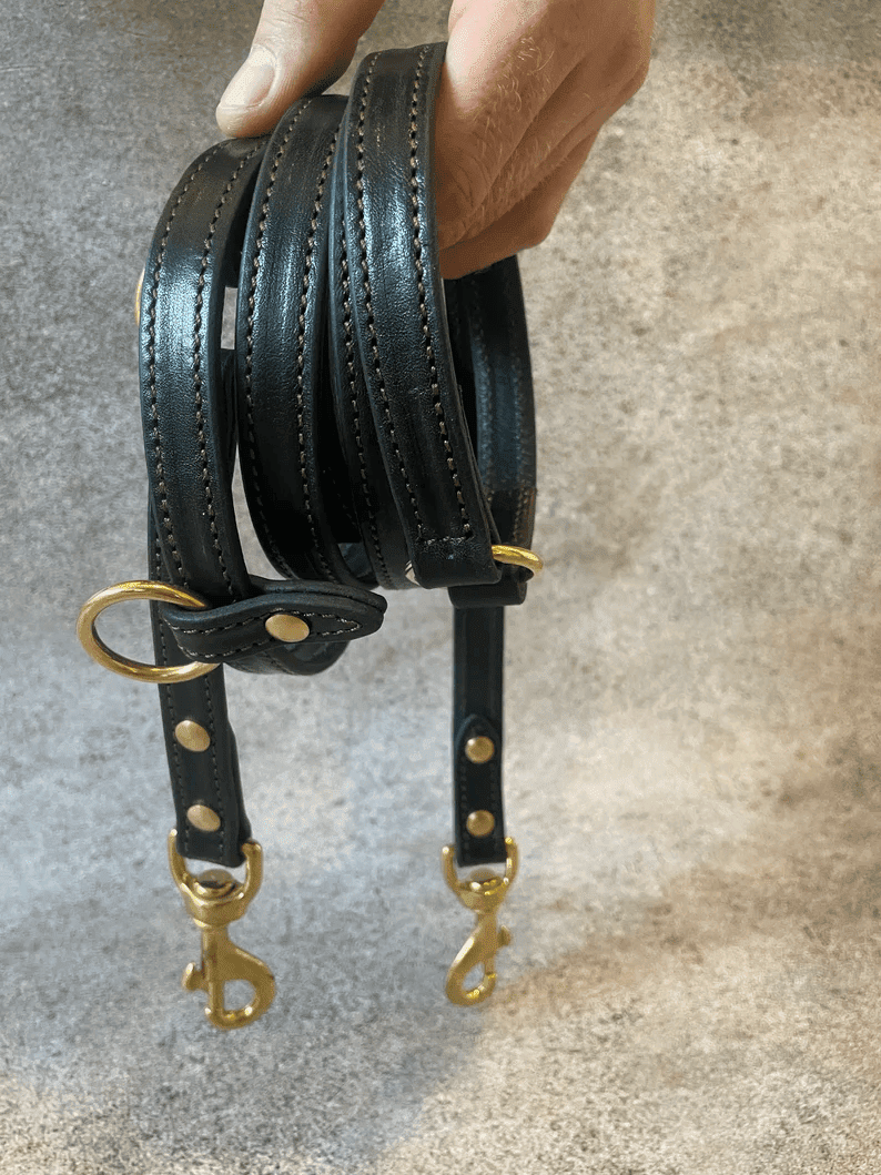 A close-up shot of a person holding a coiled, dark brown leather multi-purpose dog leash. The image highlights the detailed brown stitching, brass rivets, and two heavy-duty brass snap hooks hanging at the bottom.