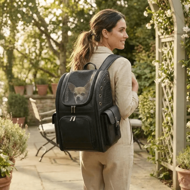 Pet Backpack Carrier | Genuine Leather A woman walking through a garden wearing the black leather Pet Backpack Carrier on her back. A small dog is visible through the mesh window, illustrating the carrier's practical use and stylish aesthetic for pet owners on the go.