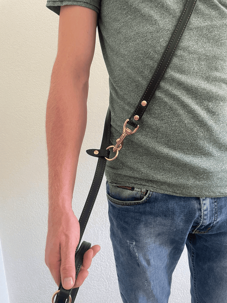 A side view of a person wearing the leather dog leash across their torso like a crossbody bag. The strap goes from the shoulder to the opposite hip, where it is secured with a brass clip and O-ring, demonstrating hands-free utility.
