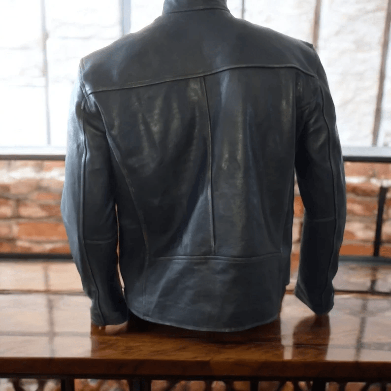 The back view of a dark blue or charcoal distressed leather jacket displayed on a mannequin. The jacket shows a simple, structured design with a horizontal seam across the upper back and vertical seams running down toward the hem.