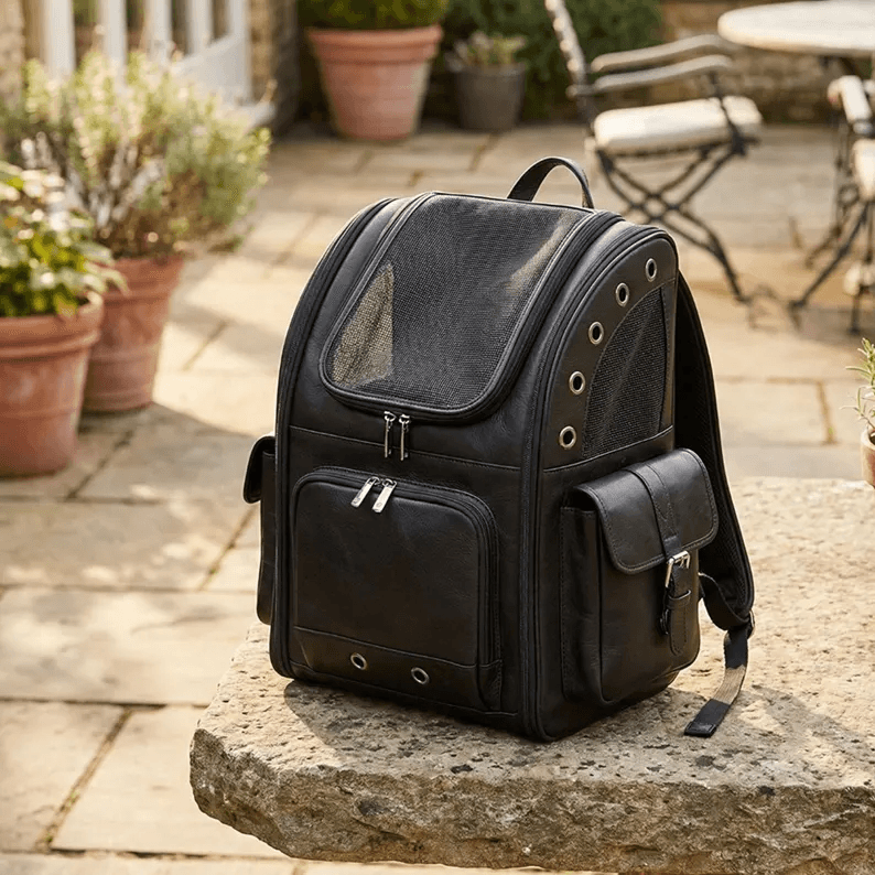 Pet Backpack Carrier | Genuine Leather A front-facing shot of the black leather Pet Backpack Carrier resting on a natural stone surface in a garden. The image highlights the large mesh window, dual front zippers, and multiple silver-toned ventilation grommets on the sides.