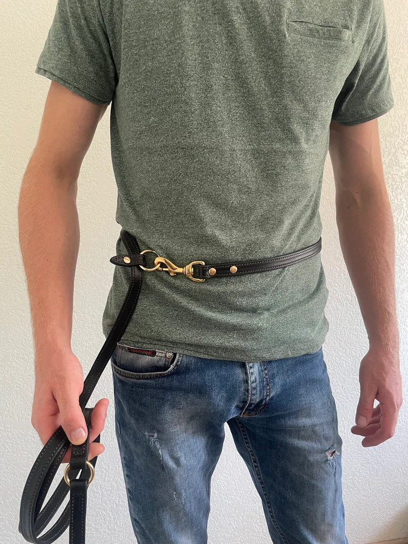Multi Purpose Dog Leash | Dog Leash Leather 8 ft A person wearing a dark leather leash configured as a hands-free waist belt. One end of the leash is clipped to a brass O-ring at the hip, showing how the leather strap fits securely around the waist of a person wearing jeans and a green t-shirt.