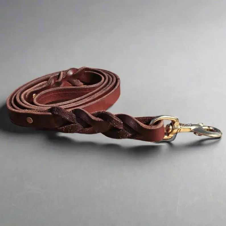 A coiled deep burgundy Latigo leather dog leash on a gray surface, highlighting the hand-braided detail near the solid brass bolt snap.