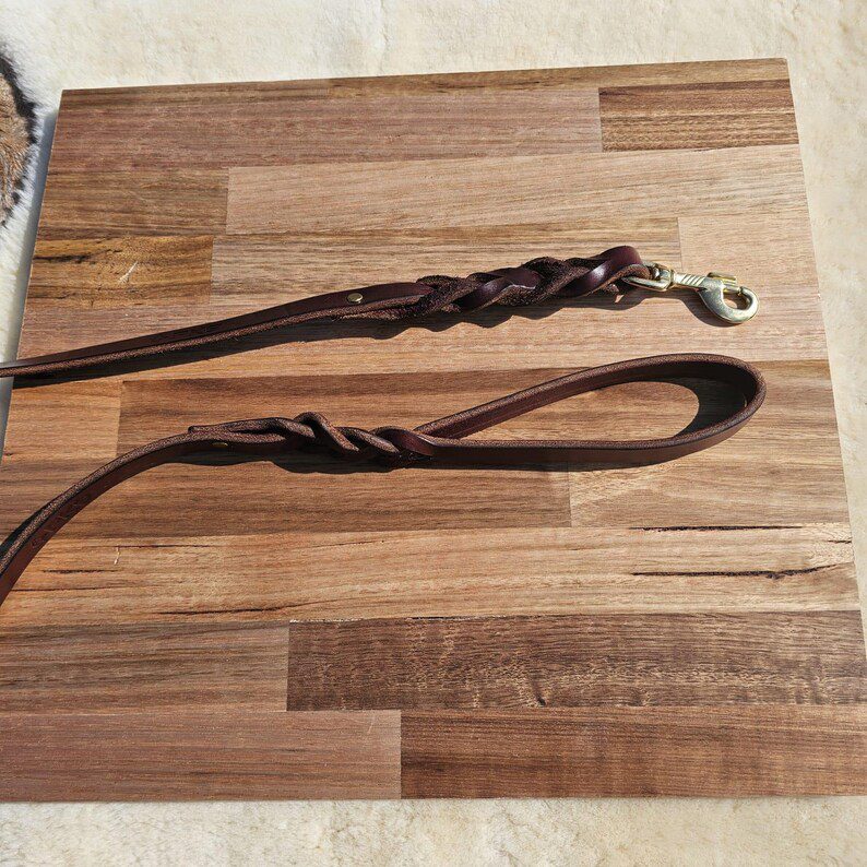 A Latigo leather dog leash laid out on a wooden surface, showcasing the loop handle with a decorative twist and the heavy-duty brass clasp at the opposite end.