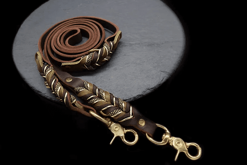 A high-quality brown leather dog leash coiled on a circular dark slate stone. The leash features intricate hand-braided details with gold-tone accents and heavy-duty brass swivel snap hooks.