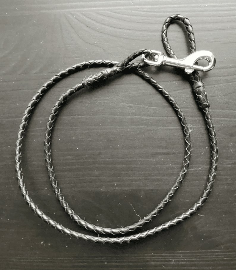 Leather Dog Leash | Braided Kangaroo Leather Dog Show Lead A full view of a slim, black braided leather dog leash coiled on a dark wood surface. The leash features a silver swivel bolt snap clasp and a small integrated loop handle.