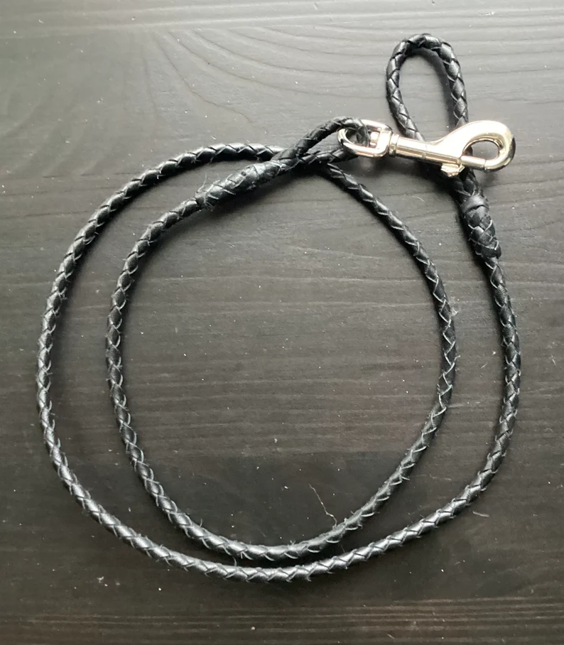Leather Dog Leash | Braided Kangaroo Leather Dog Show Lead A full view of a slim, black braided leather dog leash coiled on a dark wood surface. The leash features a silver swivel bolt snap clasp and a small integrated loop handle.
