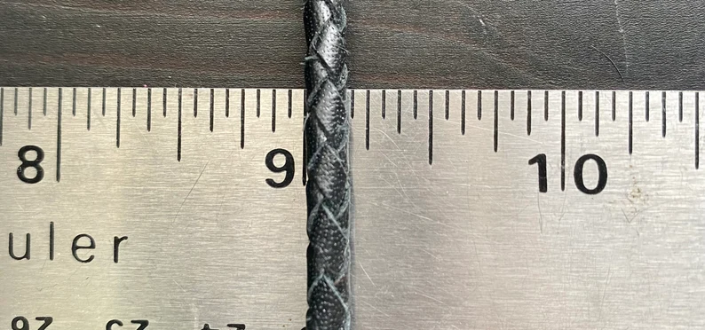 Leather Dog Leash | Braided Kangaroo Leather Dog Show Lead A macro shot of the black braided leather cord placed vertically against a metal ruler to show its thickness. The round braid measures approximately 1/4 inch in diameter.