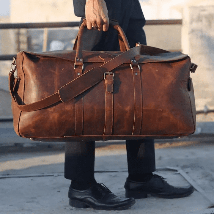 Full Grain Leather Duffel Bag | Genuine Leather Travel Bag A person dressed in professional attire holds a large, vintage-style brown leather duffel bag by its reinforced top handles while standing outdoors.