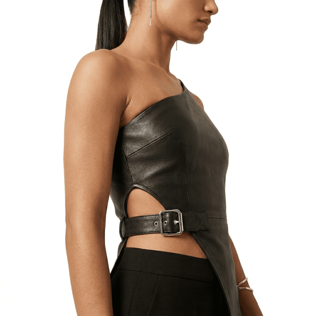 Asymmetrical One Shoulder Top | Leather One Shoulder Top A close-up, side-profile view focusing on the midsection and shoulder of a woman wearing a black leather asymmetrical one-shoulder top. The image highlights a prominent silver buckle fastening a thick leather belt over an architectural side cutout at the waist, showing a glimpse of skin. She is paired with tailored black trousers.